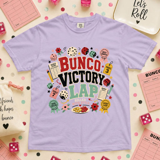 A light purple crewneck t-shirt with a colorful 'BUNCO VICTORY LAP' graphic print, styled in a flat-lay arrangement with pink bingo cards, dice, and a decorative tray on a polka-dotted background.