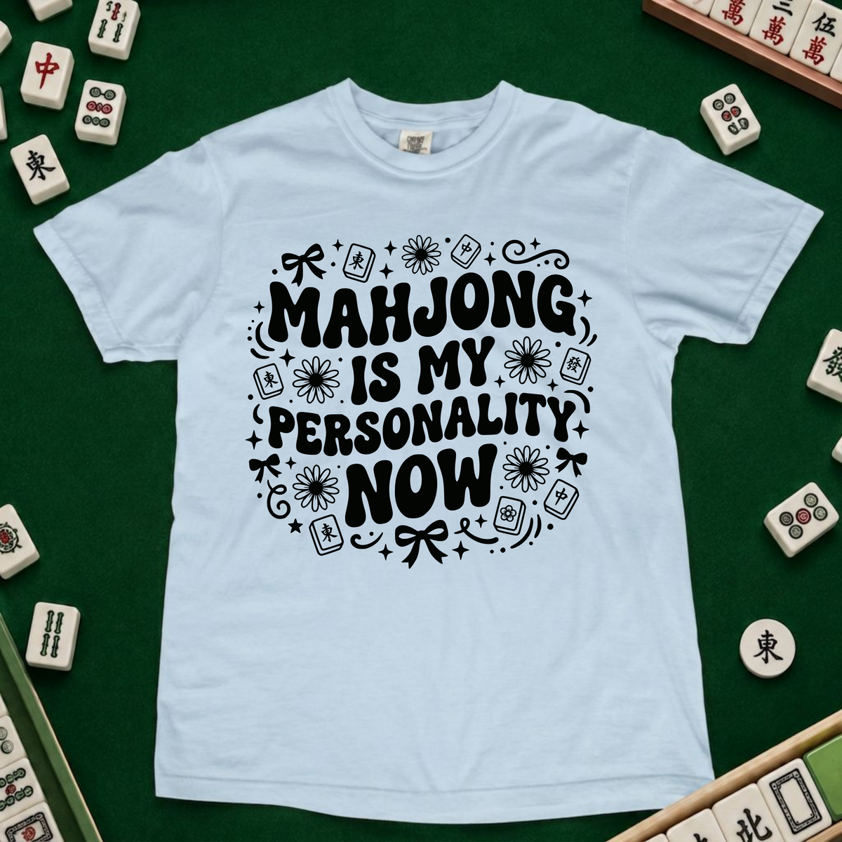 A light blue crewneck t-shirt with a black graphic text reading 'MAHJONG IS MY PERSONALITY NOW' surrounded by mahjong tile illustrations, displayed on a green felt surface with real mahjong tiles arranged around it.