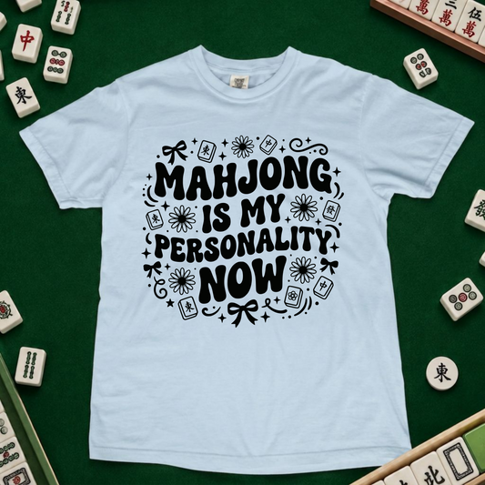 A light blue crewneck t-shirt with a black graphic text reading 'MAHJONG IS MY PERSONALITY NOW' surrounded by mahjong tile illustrations, displayed on a green felt surface with real mahjong tiles arranged around it.