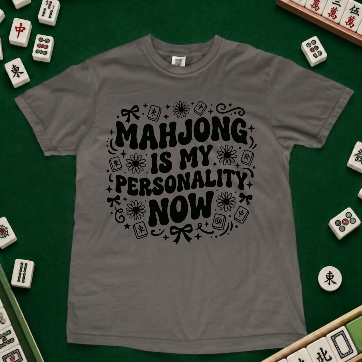 A gray short-sleeve t-shirt with a black graphic print reading 'MAHJONG IS MY PERSONALITY NOW' surrounded by mahjong-themed illustrations, laid flat on a green felt surface with mahjong tiles scattered around.