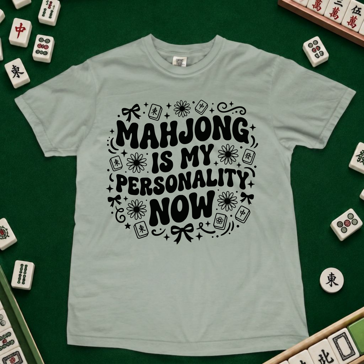 A light sage green t-shirt with a black graphic text reading 'MAHJONG IS MY PERSONALITY NOW' surrounded by mahjong tile illustrations, laid flat on a green felt surface with real mahjong tiles visible around the edges.