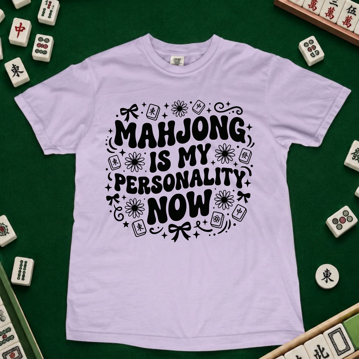 A light lavender crewneck t-shirt with a black graphic text reading 'MAHJONG IS MY PERSONALITY NOW' surrounded by mahjong tile illustrations, laid flat on a green felt surface with actual mahjong tiles arranged around it.