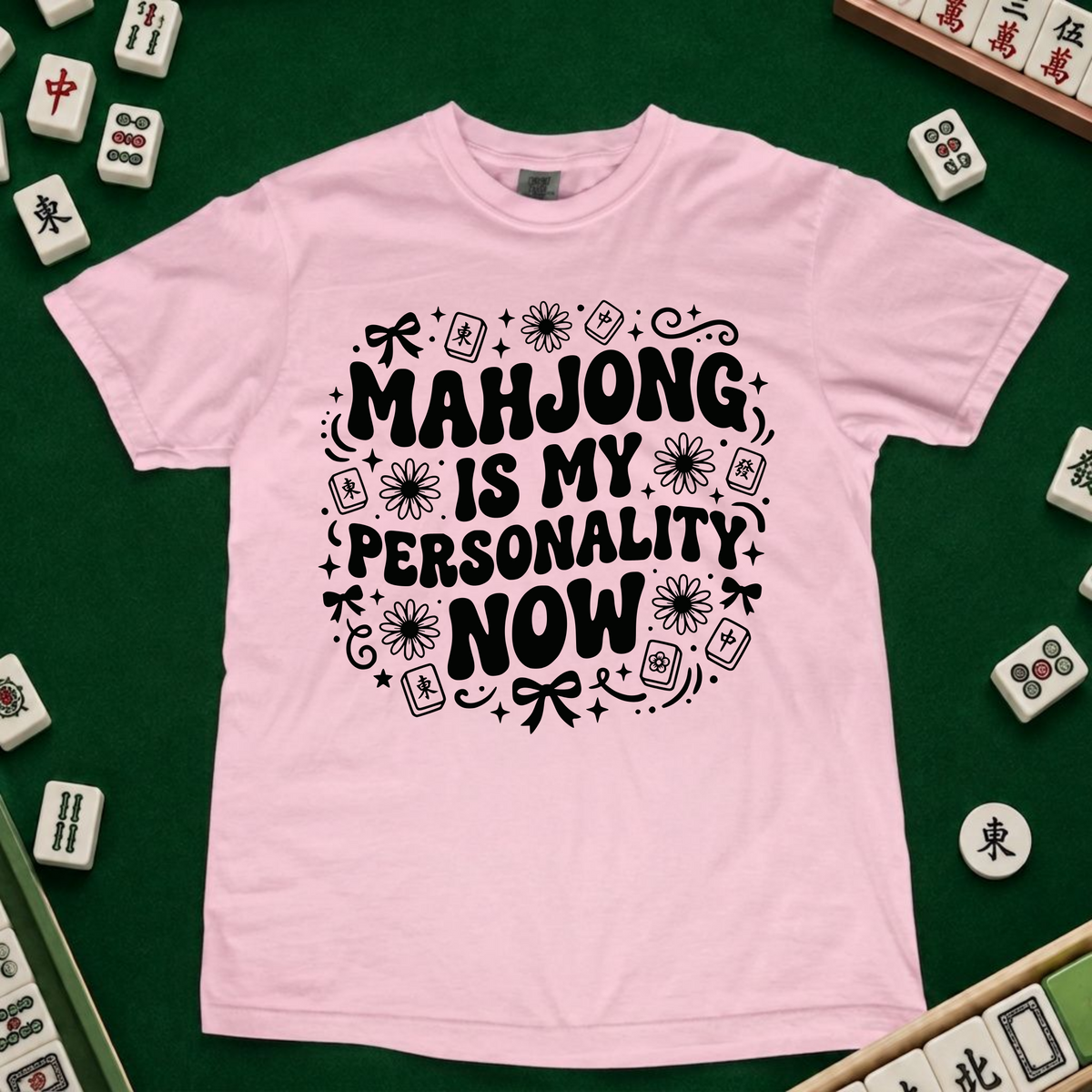 A light lavender crewneck t-shirt with a black graphic text reading 'MAHJONG IS MY PERSONALITY NOW' surrounded by mahjong tile illustrations, laid flat on a green felt surface with actual mahjong tiles arranged around it.
