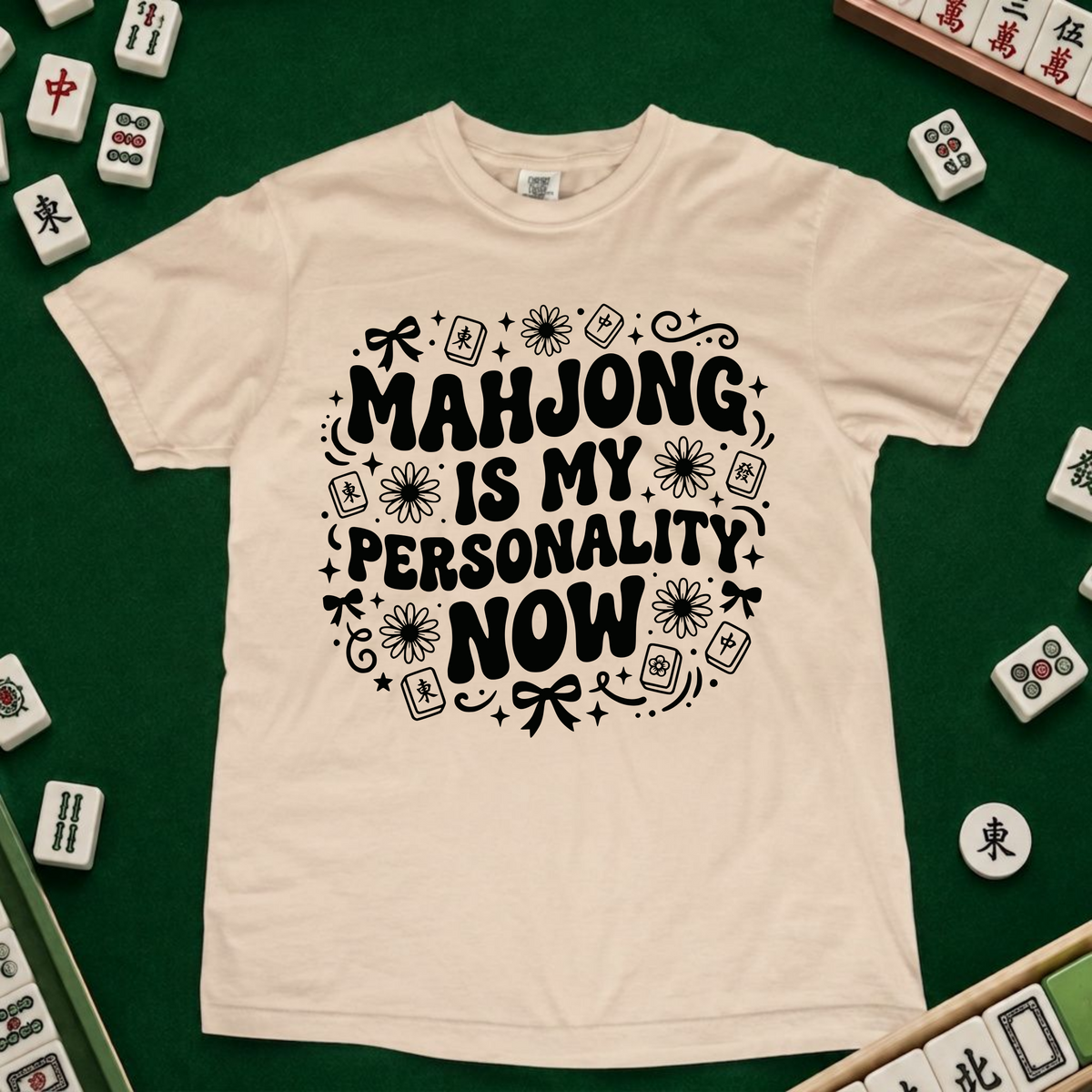 A cream-colored t-shirt with a black graphic print reading 'MAHJONG IS MY PERSONALITY NOW' surrounded by mahjong tile illustrations, laid flat on a green felt surface with scattered mahjong tiles around it.