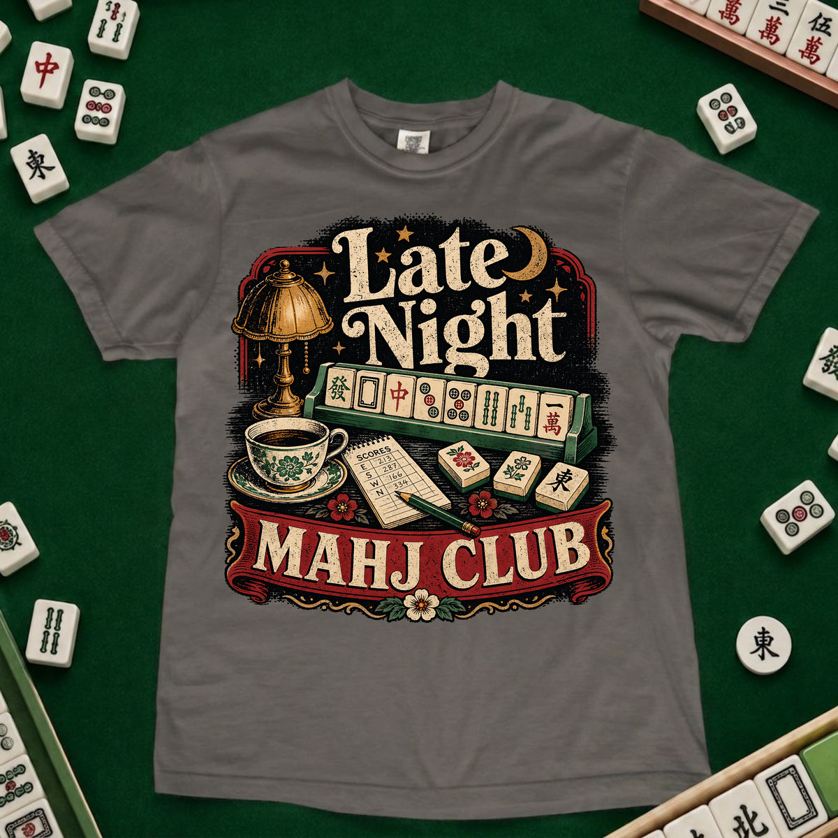 A gray short-sleeve t-shirt with a vintage-style graphic reading 'Late Night MAHJ CLUB' over a mahjong scene, laid flat on a green felt surface with real mahjong tiles visible around the edges.
