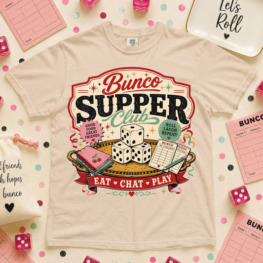 A beige graphic t-shirt featuring a 'Bunco Supper Club' design, styled in a flat-lay arrangement with pink dice, notepads, and other party accessories on a white background with pink polka dots.