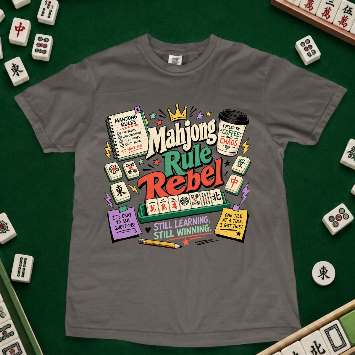 A gray short-sleeve t-shirt with a colorful 'Mahjong Rule Rebel' graphic print, laid flat on a green felt surface surrounded by miniature mahjong tiles.