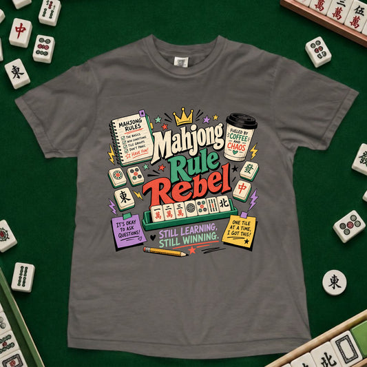 A gray short-sleeve t-shirt with a colorful 'Mahjong Rule Rebel' graphic print, laid flat on a green felt surface surrounded by miniature mahjong tiles.