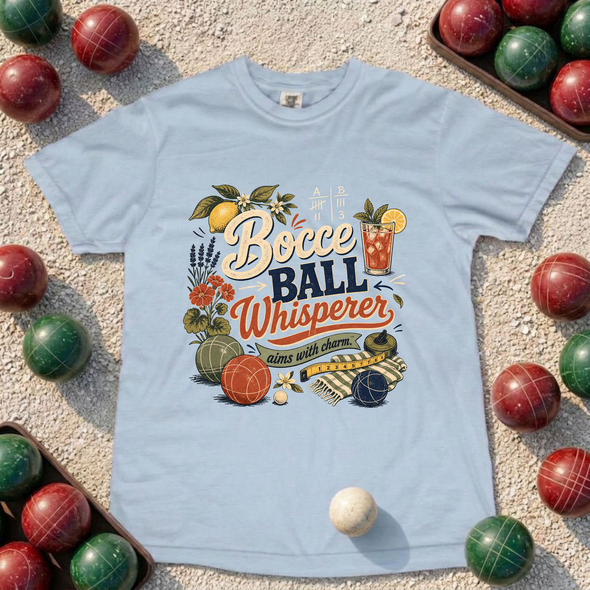 A light blue graphic t-shirt laid flat on a sandy surface, surrounded by red and green wooden bocce balls. The shirt features a colorful retro-style graphic with the text 'Bocce BALL Whisper' and 'aim with charm' surrounded by illustrations of bocce balls, lemons, and a drink.