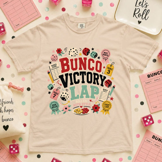 A beige graphic t-shirt featuring a colorful 'BUNCO VICTORY LAP' design, styled in a flat lay with pink dice, checklists, and polka dot accents on a white background.