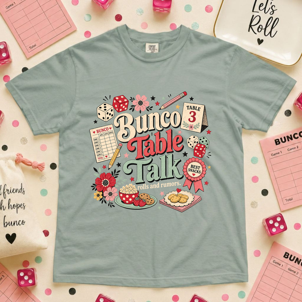 A light blue-green graphic t-shirt featuring a 'Bunco Table Talk' design with illustrations of dice, flowers, and a cake, laid flat on a pink polka-dot background with party accessories.