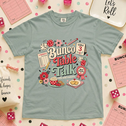 A light blue-green graphic t-shirt featuring a 'Bunco Table Talk' design with illustrations of dice, flowers, and a cake, laid flat on a pink polka-dot background with party accessories.