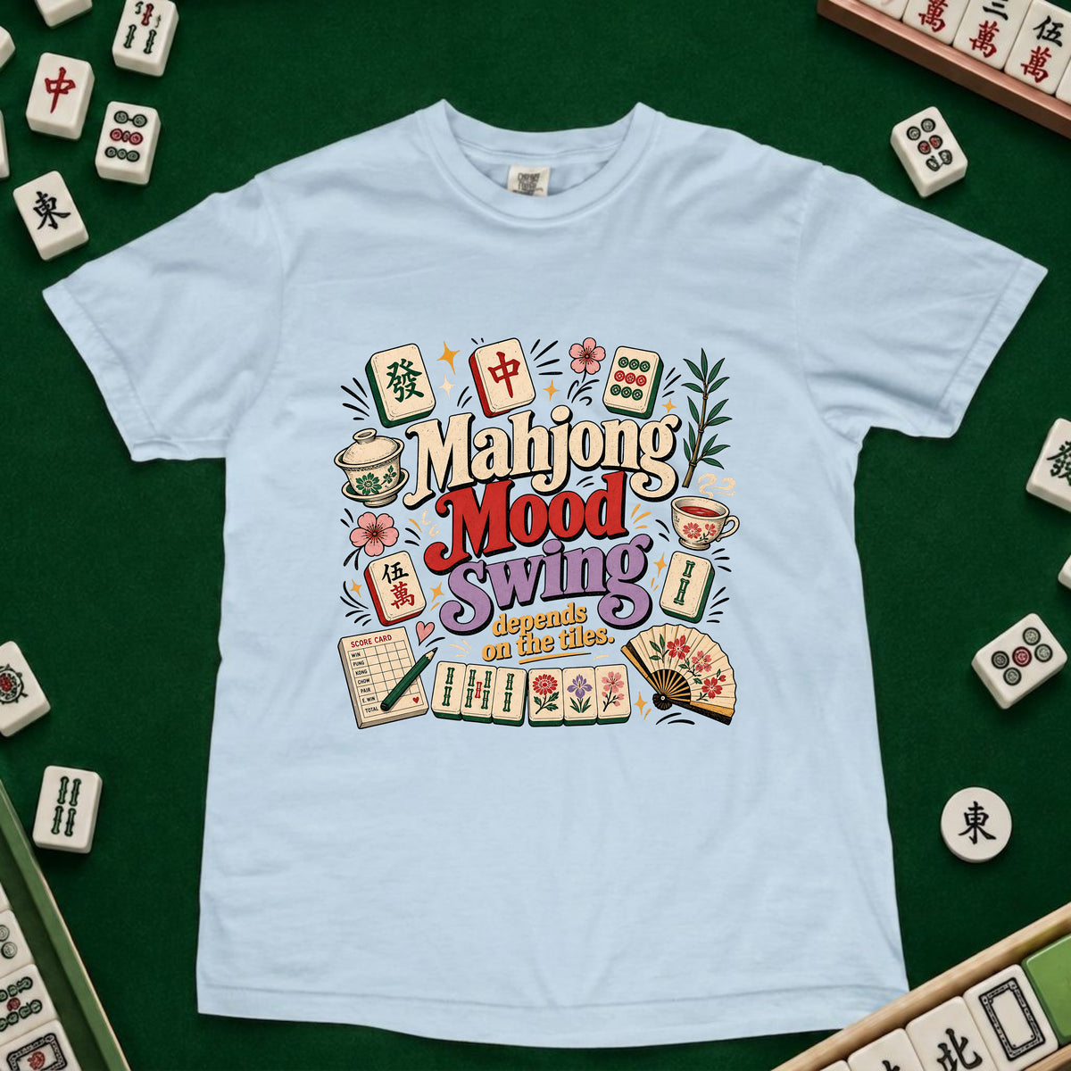 A light blue crewneck t-shirt featuring a colorful graphic design with mahjong tiles, cups, and the text 'Mahjong Mood Swing depends on the tiles' laid out on a green felt surface surrounded by real mahjong tiles.