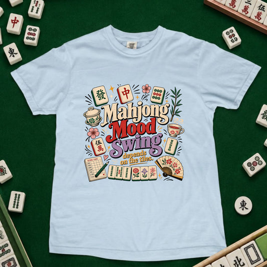A light blue crewneck t-shirt featuring a colorful graphic design with mahjong tiles, cups, and the text 'Mahjong Mood Swing depends on the tiles' laid out on a green felt surface surrounded by real mahjong tiles.