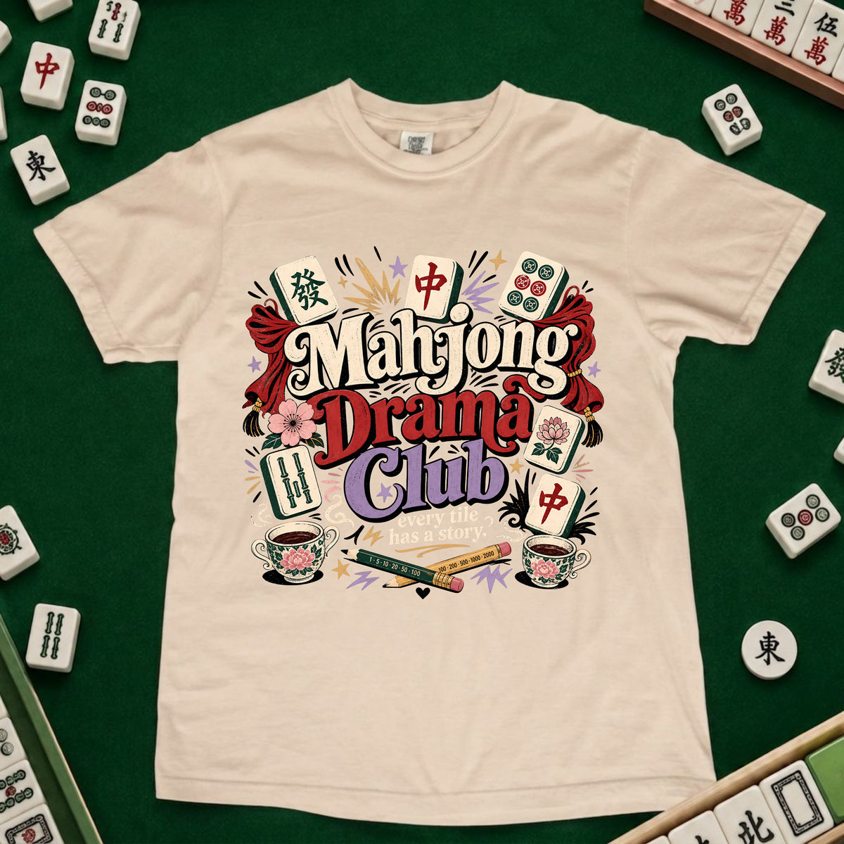 A cream-colored short-sleeve t-shirt featuring a colorful graphic print that reads 'Mahjong Drama Club' surrounded by illustrations of mahjong tiles, cups, and cards. The shirt is laid flat on a green felt surface, with real mahjong tiles arranged around the edges as props.