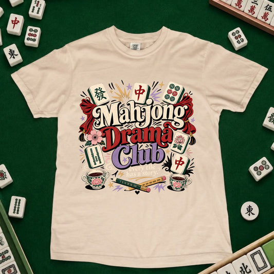 A cream-colored short-sleeve t-shirt featuring a colorful graphic print that reads 'Mahjong Drama Club' surrounded by illustrations of mahjong tiles, cups, and cards. The shirt is laid flat on a green felt surface, with real mahjong tiles arranged around the edges as props.