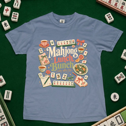 A light blue crewneck t-shirt featuring a colorful retro graphic that reads 'Mahjong Lunch Bunch' surrounded by illustrations of tea cups, food, and mahjong tiles. The shirt is laid flat on a green felt surface, surrounded by real mahjong tiles.