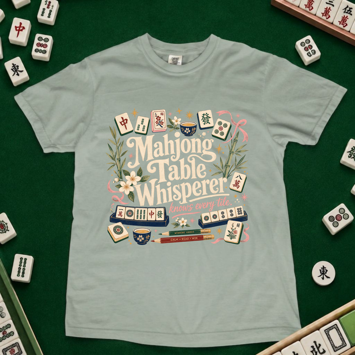 A light sage green t-shirt featuring a colorful graphic design of mahjong tiles and the words 'Mahjong Table Whisperer' surrounded by flowers and greenery, laid flat on a dark green felt surface with mahjong tiles scattered around the edges.
