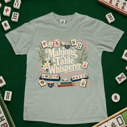 A light sage green t-shirt featuring a colorful graphic design of mahjong tiles and the words 'Mahjong Table Whisperer' surrounded by flowers and greenery, laid flat on a dark green felt surface with mahjong tiles scattered around the edges.