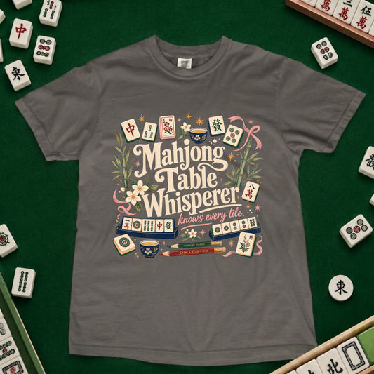 A gray short-sleeve t-shirt with a colorful graphic print reading 'Mahjong Table Whisperer' surrounded by illustrated mahjong tiles and floral details, laid flat on a green felt surface with real mahjong tiles visible around the edges.