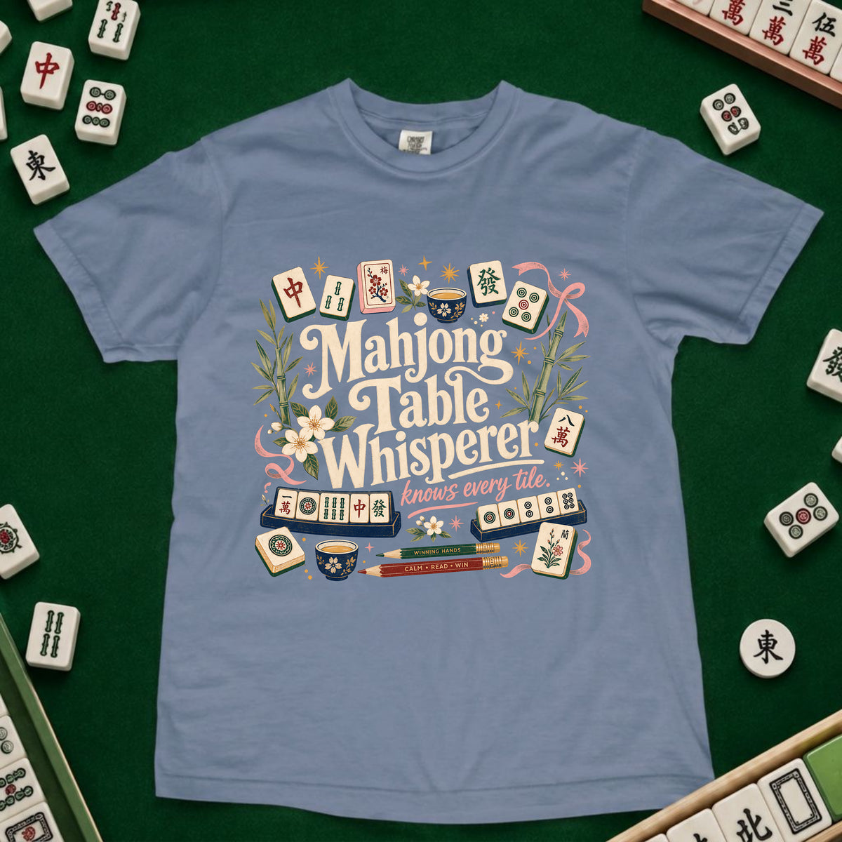 A light blue crewneck t-shirt featuring a colorful graphic design of mahjong tiles and the words 'Mahjong Table Whisperer' on a green felt background with scattered mahjong tiles.