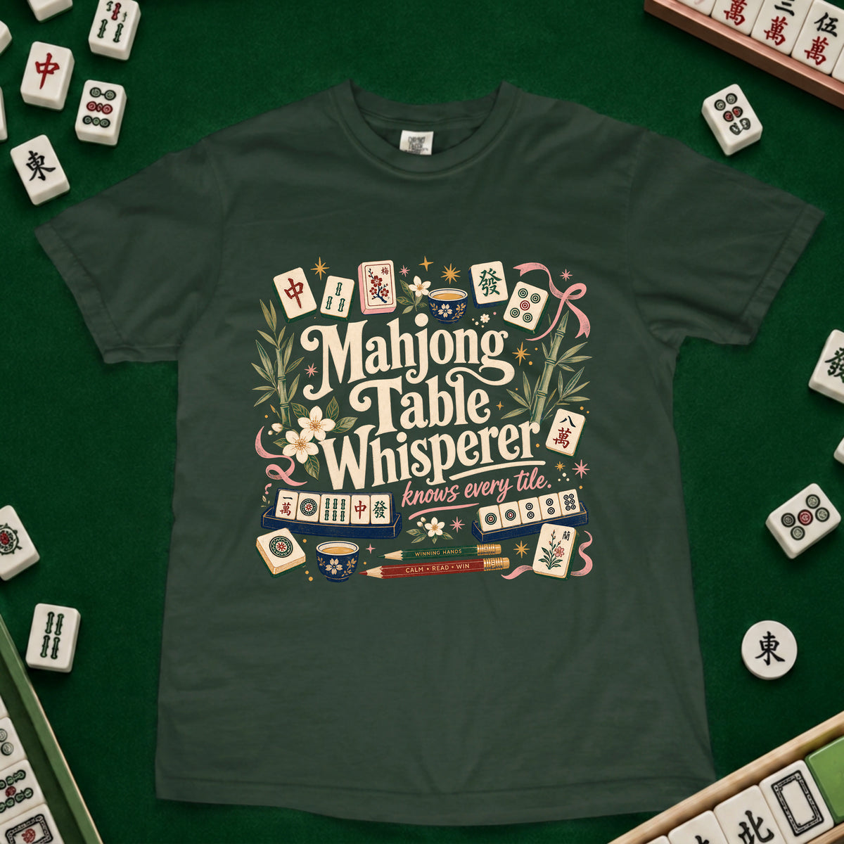 A dark green short-sleeve t-shirt featuring a colorful graphic print of mahjong tiles and the text 'Mahjong Table Whisperer knows every tile'. The shirt is laid flat on a green felt surface, surrounded by scattered real mahjong tiles.