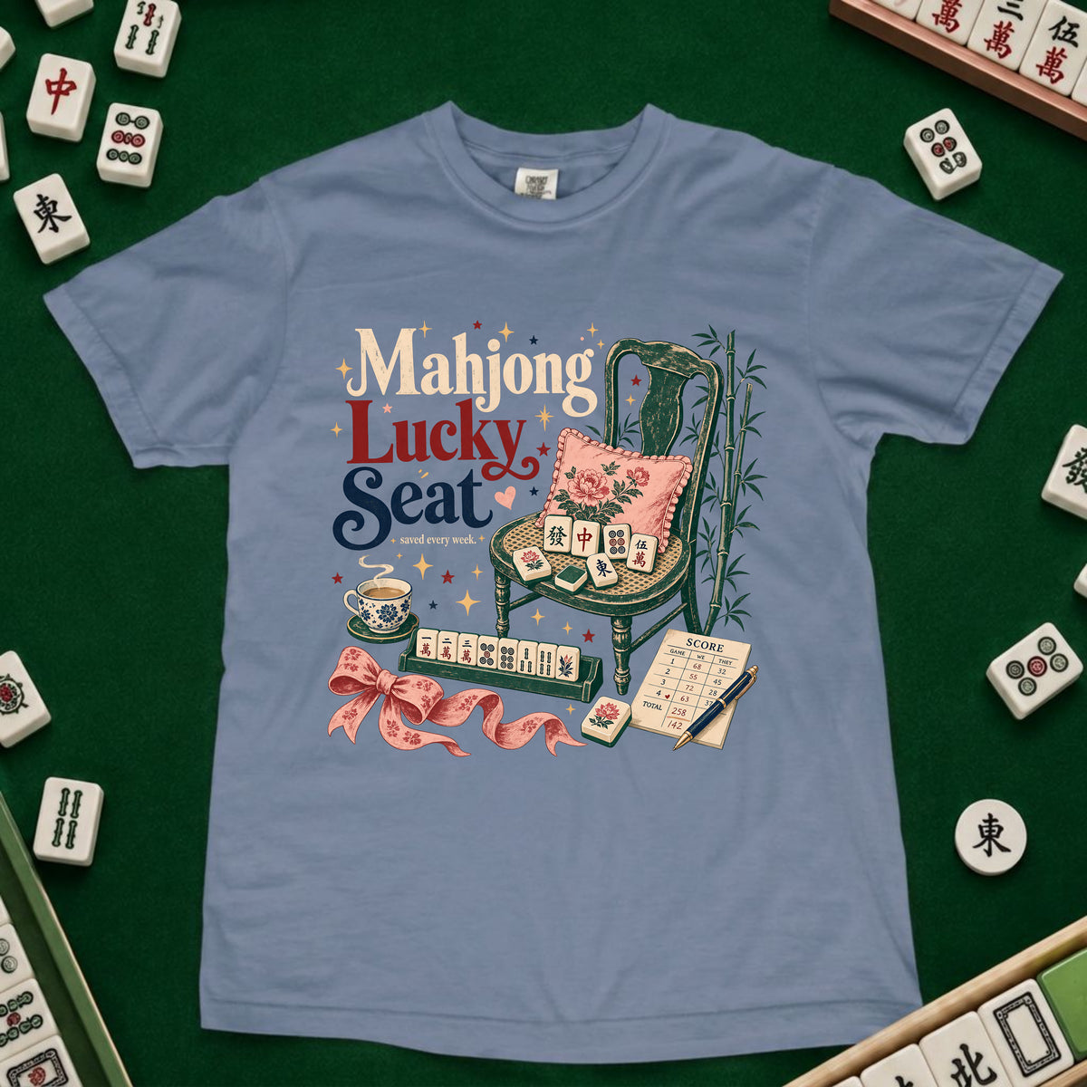 A light blue graphic t-shirt featuring a 'Mahjong Lucky Seat' design with a green chair, pink cushion, and mahjong tiles, laid flat on a green felt surface surrounded by real mahjong tiles.