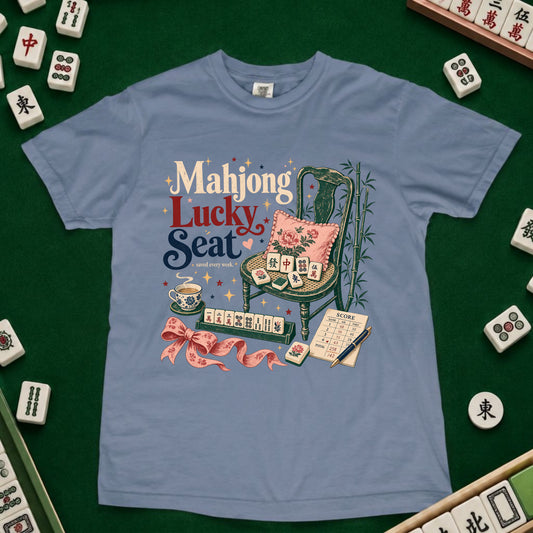 A light blue graphic t-shirt featuring a 'Mahjong Lucky Seat' design with a green chair, pink cushion, and mahjong tiles, laid flat on a green felt surface surrounded by real mahjong tiles.