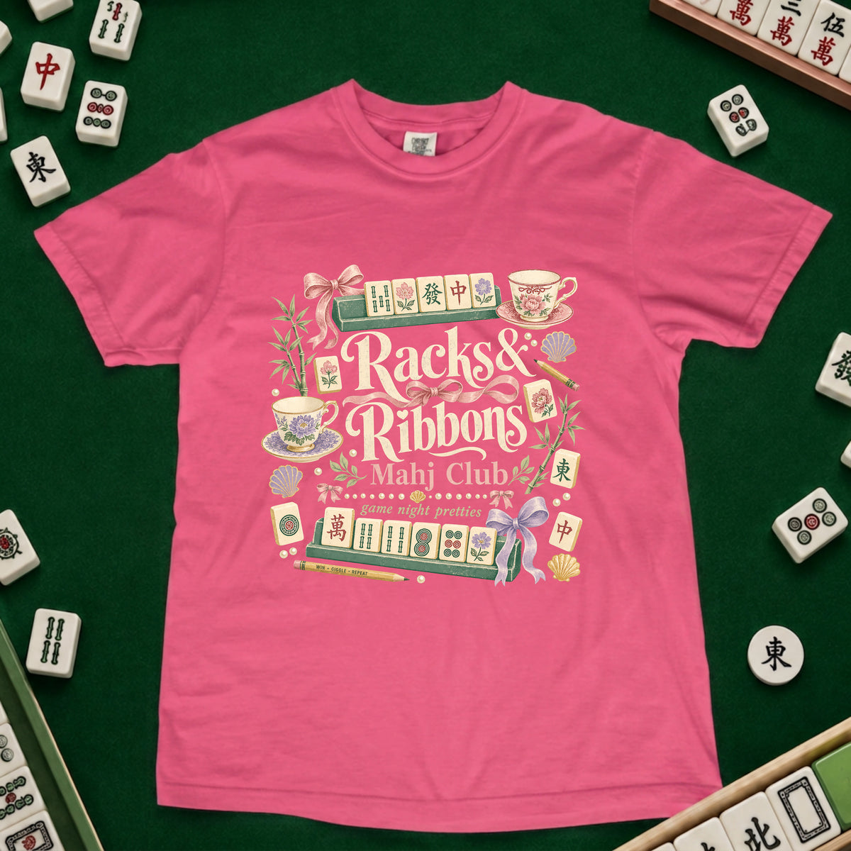 A bright pink short-sleeve t-shirt with a detailed graphic print reading 'Racks & Ribbons Mahj Club' surrounded by illustrated mahjong tiles, teacups, and ribbons, laid flat on a green felt surface with real mahjong tiles scattered around the edges.