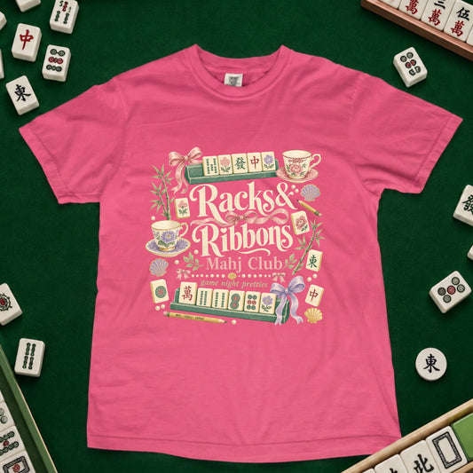 A bright pink short-sleeve t-shirt with a detailed graphic print reading 'Racks & Ribbons Mahj Club' surrounded by illustrated mahjong tiles, teacups, and ribbons, laid flat on a green felt surface with real mahjong tiles scattered around the edges.