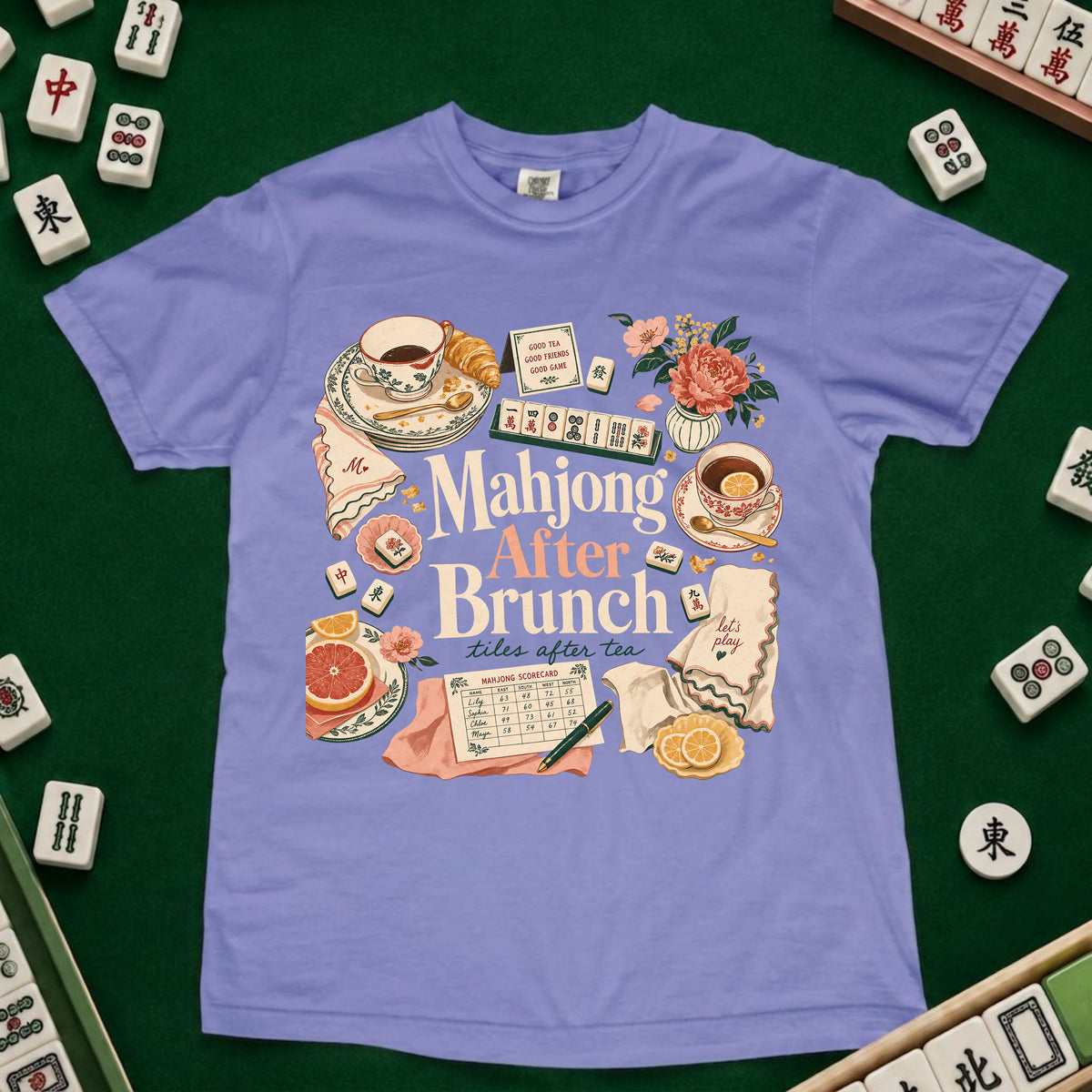A light purple short-sleeve t-shirt with a colorful graphic print reading 'Mahjong After Brunch' surrounded by tea cups, flowers, cards, and food, laid flat on a green felt surface with mahjong tiles around the edges.