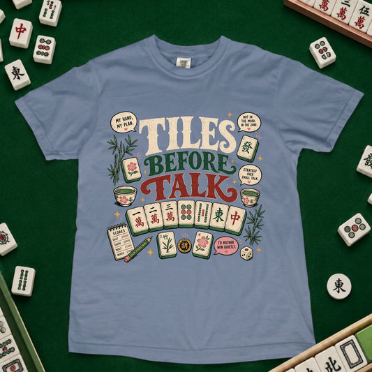A light blue crewneck t-shirt featuring a colorful graphic design with the words 'TILES BEFORE TALK' surrounded by illustrations of mahjong tiles, chips, and scoring sticks, laid flat on a green felt surface with real mahjong tiles scattered around the edges.