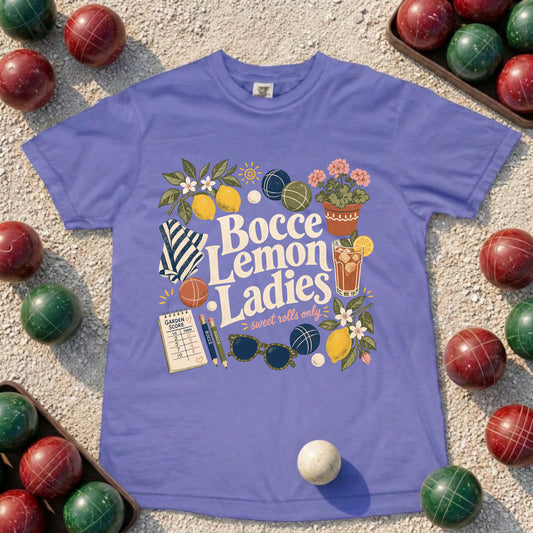 A purple short-sleeve graphic T-shirt laid flat on a sandy surface, surrounded by red and green croquet balls. The shirt features a colorful retro-style chest print reading 'Bocce Lemon Ladies' with illustrations of lemons, flowers, sunglasses, a striped tie, and sports items.