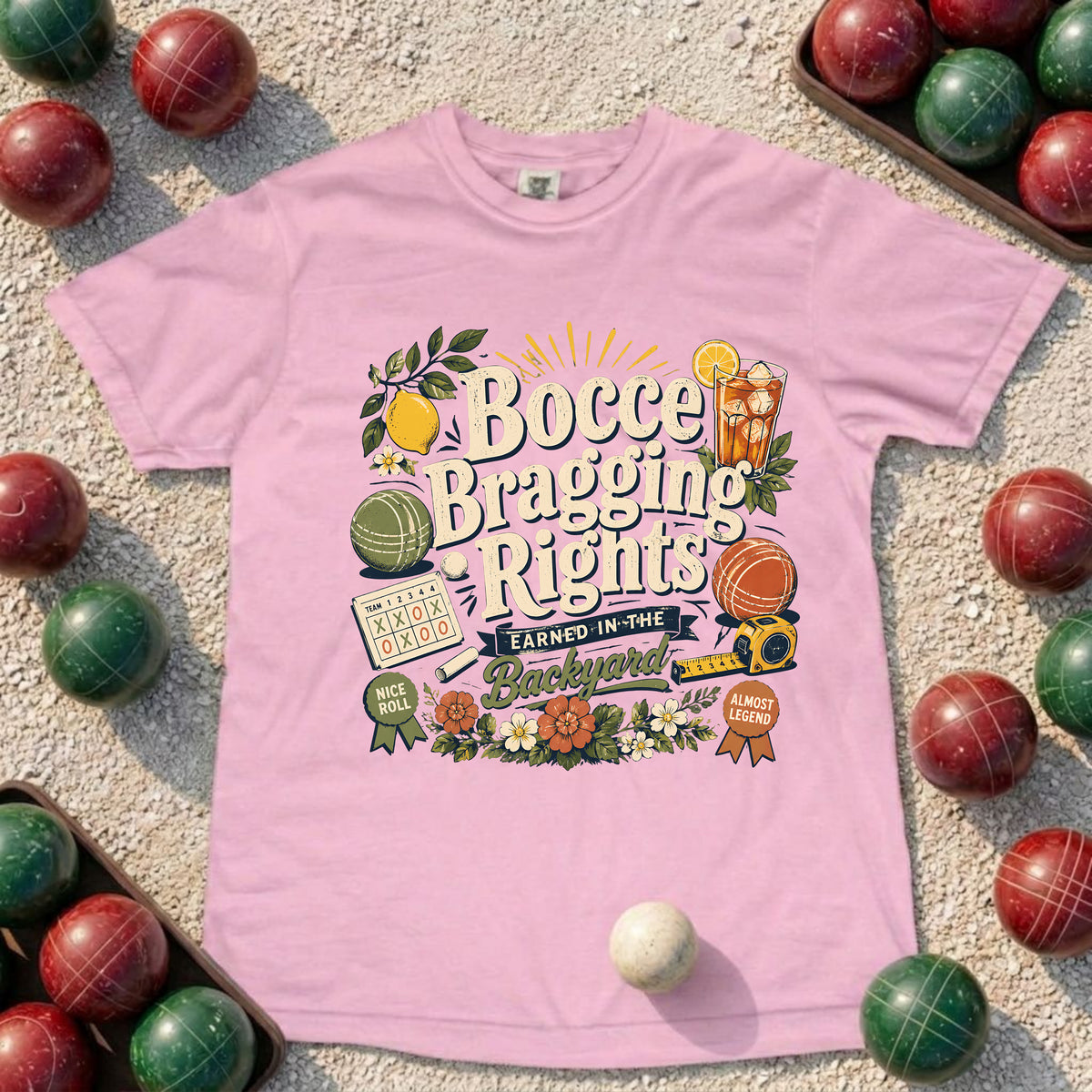 A light pink short-sleeve graphic T-shirt laid flat on a sandy surface, surrounded by red and green croquet balls. The shirt features a colorful retro-style front print reading 'Bocce Bragging Rights' with croquet-themed illustrations and badges.