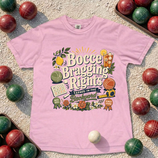 A light pink short-sleeve graphic T-shirt laid flat on a sandy surface, surrounded by red and green croquet balls. The shirt features a colorful retro-style front print reading 'Bocce Bragging Rights' with croquet-themed illustrations and badges.