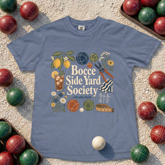 A dusty blue crewneck t-shirt laid flat on a sandy surface, surrounded by red and green bowling balls. The shirt features a colorful graphic design with the text 'Bocce Side Yard Society' and illustrations of bowling props like balls, a drink, flowers, and a bowling pin.