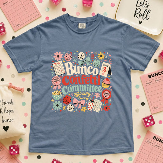 A blue crewneck t-shirt featuring a colorful 'Bunco Confetti Committee' graphic, styled in a flat-lay arrangement with pink bingo cards, dice, and confetti on a light background.