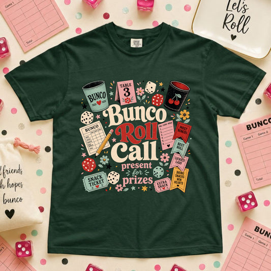 A dark green short-sleeve t-shirt with a colorful 'Bunco Roll Call' graphic print, laid flat on a light pink background decorated with dice, bingo cards, and party supplies.