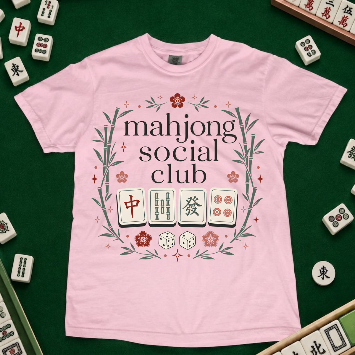 A light pink short-sleeve t-shirt with a 'mahjong social club' graphic printed on the front, laid flat on a green felt surface surrounded by miniature mahjong tiles.