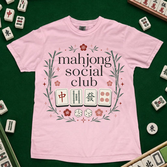 A light pink short-sleeve t-shirt with a 'mahjong social club' graphic printed on the front, laid flat on a green felt surface surrounded by miniature mahjong tiles.