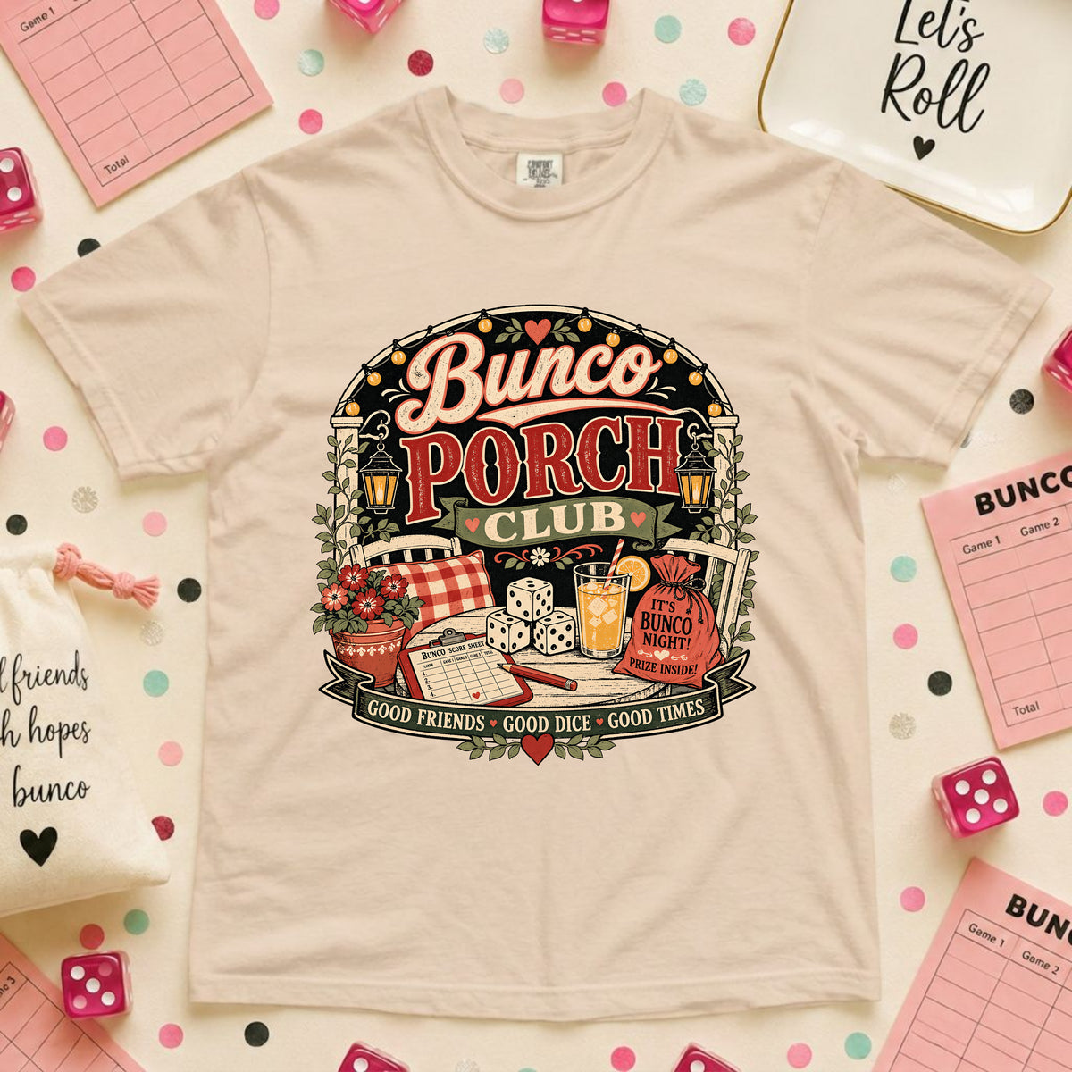 A beige graphic t-shirt featuring a 'Bunco Porch Club' design, styled in a flat-lay arrangement with pink dice, checklists, and confetti on a light background.