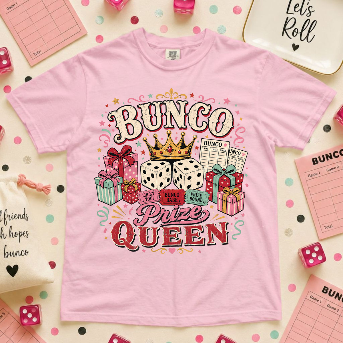 A light pink short-sleeve t-shirt with a colorful 'BUNCO Prize Queen' graphic, styled in a flat-lay arrangement with pink dice, checkered cloths, and party accessories.