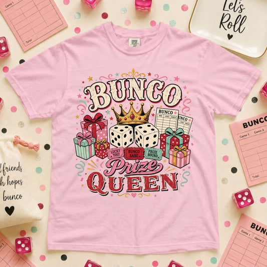 A light pink short-sleeve t-shirt with a colorful 'BUNCO Prize Queen' graphic, styled in a flat-lay arrangement with pink dice, checkered cloths, and party accessories.